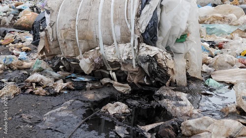Garbage is piled up at a landfill with plastic bags, waste, and debris covering the ground in an urban area during daylight hours