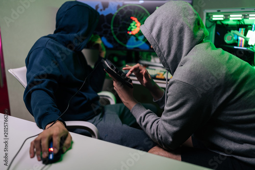 Two unrecognizable people in hooded sweatshirts sit at desk in dark room. They work on computer, focused on screen with world map and code. Green light creates mysterious, tense atmosphere.