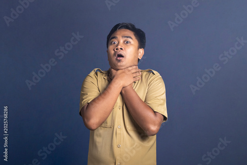 Cuadro en lienzo Panicked young man in a khaki uniform shirt looking at the camera with wide eyes and open mouth while clutching his own neck with both hands in a choking gesture against a blue background