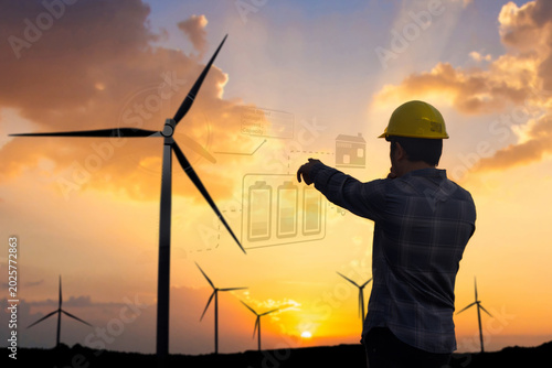 Silhouette of engineer pointing at digital hologram with battery data at wind turbine farm during sunset. Renewable energy concept.