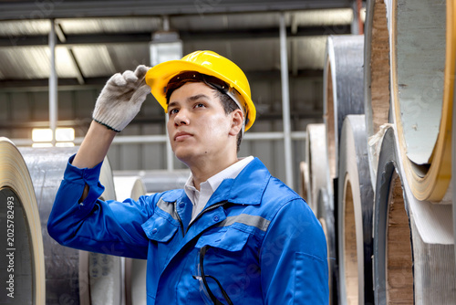 Portrait of happy young male worker wearing safety clothing and helmet, touching hard hat during inspecting large metal tube in industrial site. Inspector engineer working at manufacturing factory.