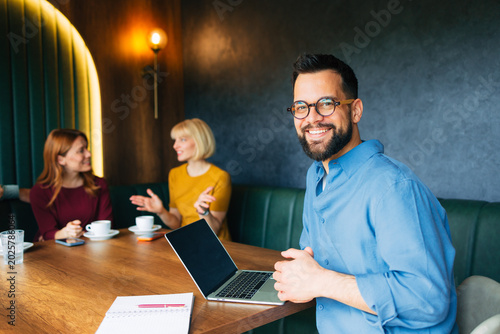 Fényképezés Confident businessman smiling at camera while sitting with laptop and coworkers