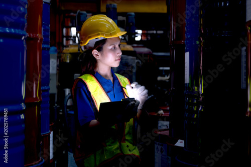 Asian female worker with tablet inspect product in large steel drums at factory during night shift. Engineer woman with helmet and safety vest work in industrial site, check product quality in a dark