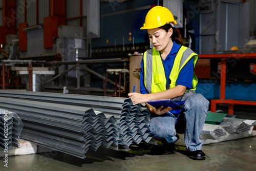 Asian female worker with tablet inspects metal sheet product at manufacturing factory during night shift. Engineer with helmet and safety vest work in industrial site, check product quality in darkใ