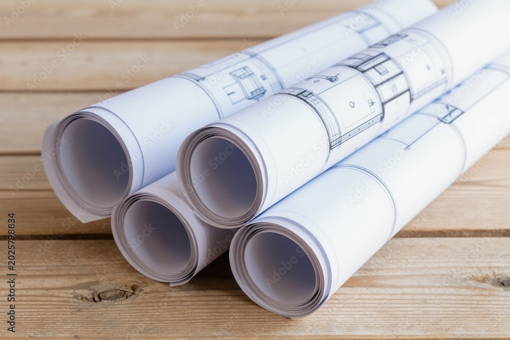 Fototapeta premium Rolled architectural blueprints lie on a light wooden table. Technical drawings display building plans and project details. Pro documents await review and work.