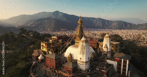 Aerial view of Swayambhunath stupa with its golden spire and colorful prayer flags glowing in warm sunset light, framed by misty Himalayan foothills.