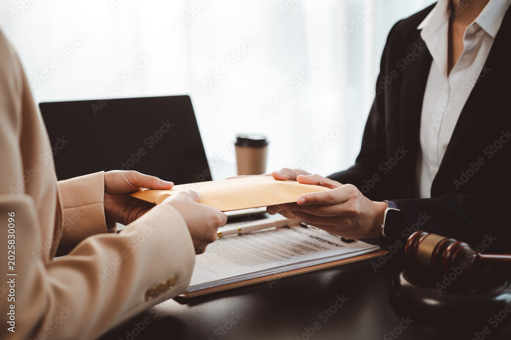 Obraz premium Businesswoman handing brown envelope containing document or money to a lawyer during a legal consultation, discussing contract agreement, corruption, or bribery with a gavel on the table
