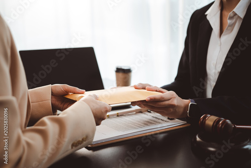 Businesswoman handing brown envelope containing document or money to a lawyer during a legal consultation, discussing contract agreement, corruption, or bribery with a gavel on the table