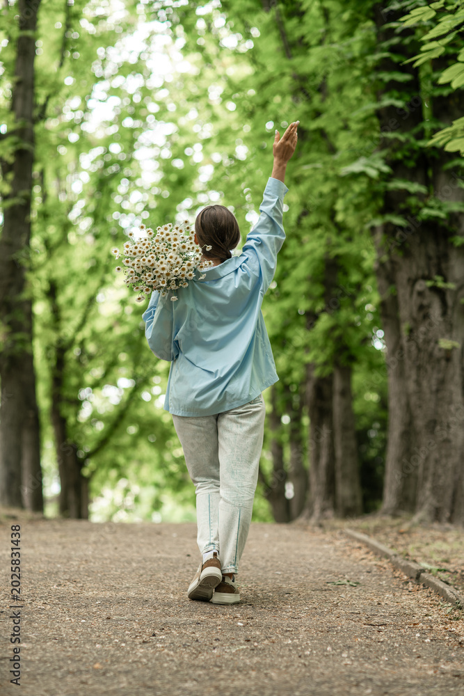 Fototapeta premium Flowers Daisies Woman Walking, Park: Woman with daisies strolls in park, raising arm on sunny day in natural setting.