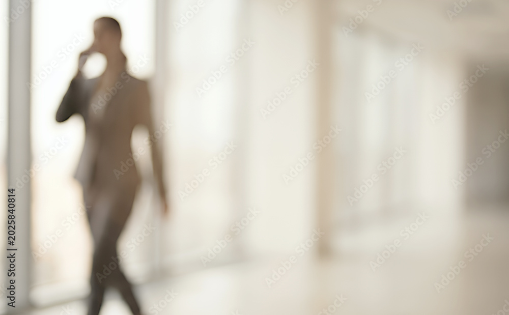 Fototapeta premium A blurred image of a businesswoman in a suit, walking confidently while using a smartphone. The setting is an office with large windows, creating a professional atmosphere.