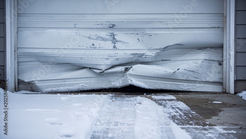 A damaged white garage door with ice and snow accumulation in a winter setting, highlighting the need for property management and maintenance to address weather-related damage.