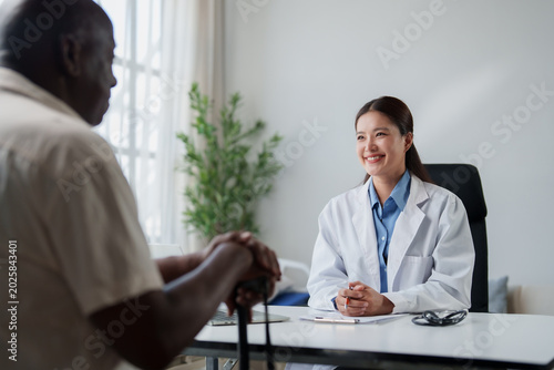 Young Asian woman doctor happily consulting with an older Black man patient, discussing his health and offering empathetic care in a professional healthcare setting