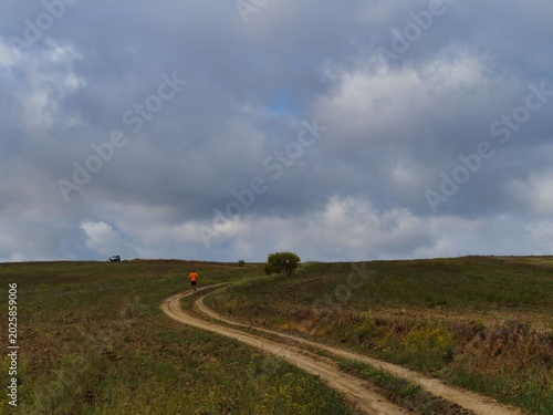 Person walks along a dirt path through hills under clouds in a rural setting during daylight hours