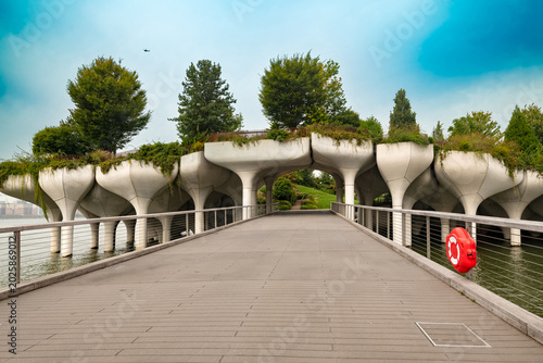 People walk along the bridge leading to Little Island at Pier 55 in Chelsea, an innovative public park featuring unique concrete tulip structures and lush green landscapes in New York City.