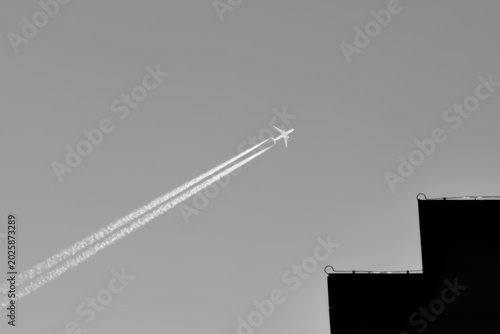 фотографія Rising to the sky: Black and white conceptual image of an airplane with a long contrail ascending over a stepped building roof silhouette