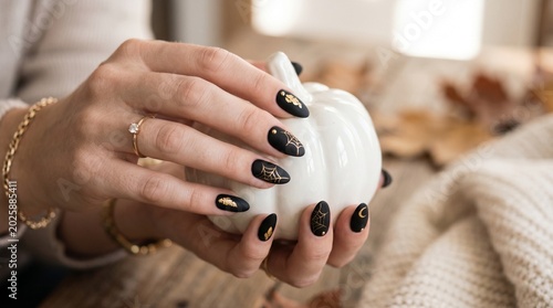 Woman's elegant hand with matte black and gold Halloween nail art holding a mini white porcelain pumpkin. Luxury cosmetic advertising scene with vast dark charcoal copy space on the right.