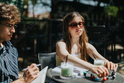 Photos A young woman and a male classmate sit at an outdoor cafe table with notebooks and drinks