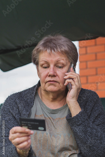 Senior woman talking on smartphone while holding bank card outdoors, concerned elderly person facing telephone fraud scam, financial security risk, cybercrime awareness consumer protection 