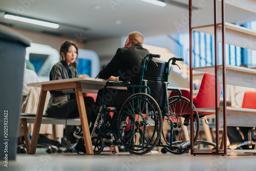 A business meeting with a man in a wheelchair speaking to a colleague at a modern office table. Colleagues sit and communicate in an accessible, collaborative workplace setting.