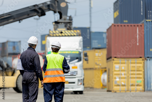 Photos Two workers in safety gear oversee container loading at a shipping yard with stacked colorful cargo containers and a truck in the background