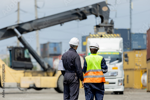 Photos Two workers in safety gear oversee a crane lifting a container at an industrial shipping yard