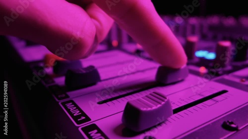 Close up of an audio mixing console featuring channel faders in a studio. The professional soundboard is illuminated by blue and red LED lights. The sliders adjust volume levels.