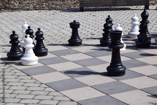  Large outdoor chess set with giant black and white pieces on a stone checkered board in a public park on a sunny day