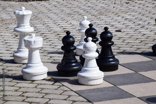  Large outdoor chess set with giant black and white pieces on a stone checkered board in a public park on a sunny day