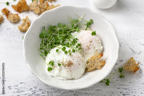 Poached eggs with microgreens and bread croutons in white bowl on light rustic background