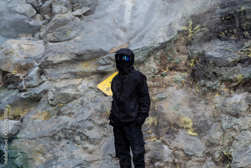 Fotografie Adventurer wearing black outdoor jacket, balaclava, and goggles standing in front of smoking volcanic rocks with sulfur deposits