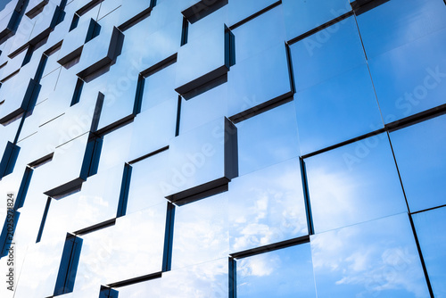 Contemporary glass facade of a commercial building featuring repeating geometric windows and reflective panels that mirror the blue sky, creating an abstract urban pattern and minimalist architecture