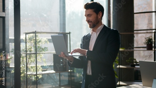 Confident Caucasian businessman typing laptop smiling in office morning sunlight professional male leader business man guy entrepreneur manager working online executive boss looking at camera indoors