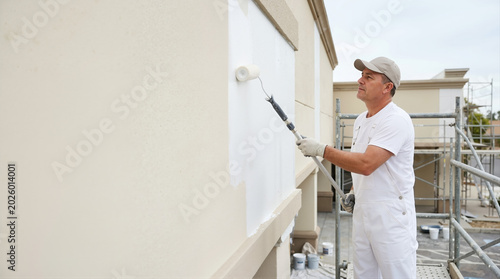 Male worker painting building facade with a roller. Professional painter in white overalls standing on scaffolding applying white paint to exterior wall. Construction and renovation concept