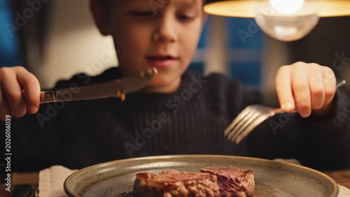 Cheerful child eating steak with knife and fork at dinner table