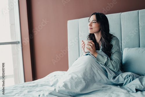 Tableau sur toile Young woman enjoying a peaceful morning coffee in bed, looking out the window an