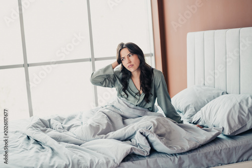 Bild auf Leinwand Tired woman sitting up in bed in morning light, rubbing her head and looking tho