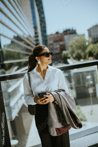 Woman stands outdoors in city while checking phone and holding a jacket