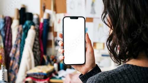 Hand holding mobile phone with blank white screen mockup, fashion designer browsing fabric collection in creative studio, clothing design and tailoring concept.
