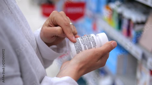 Close up of a healthcare professional's hands carefully reading the instructions and ingredients on a white medicine bottle while standing in a pharmacy aisle with blurred shelves in the background