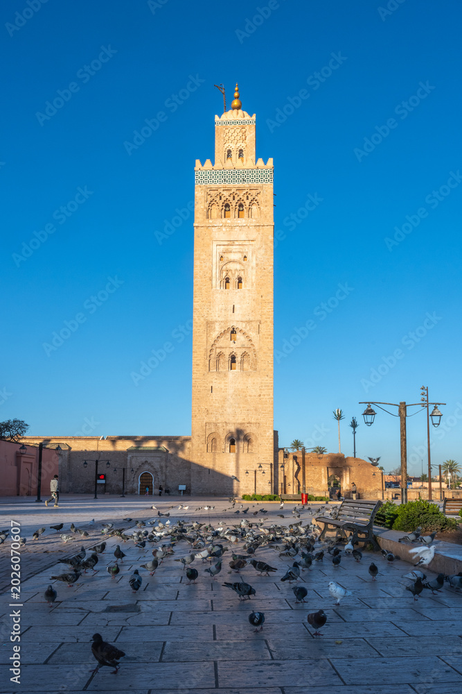Fototapeta premium Kutubiyya Mosque in Marrakesh shows its tall minaret under a clear blue sky. Pigeons gather on the ground, and visitors enjoy the space around the mosque.