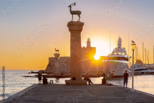 Sunrise view of Mandraki Harbor in Rhodes with deer statues, yachts and calm sea water near the historic waterfront.