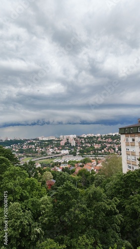 A dark storm cloud hanging over a city with buildings and forest in the background. Dramatic weather scene with contrast between urban area and nature.