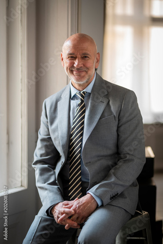 Smiling mature businessman in a classic grey suit standing by a window, looking at camera