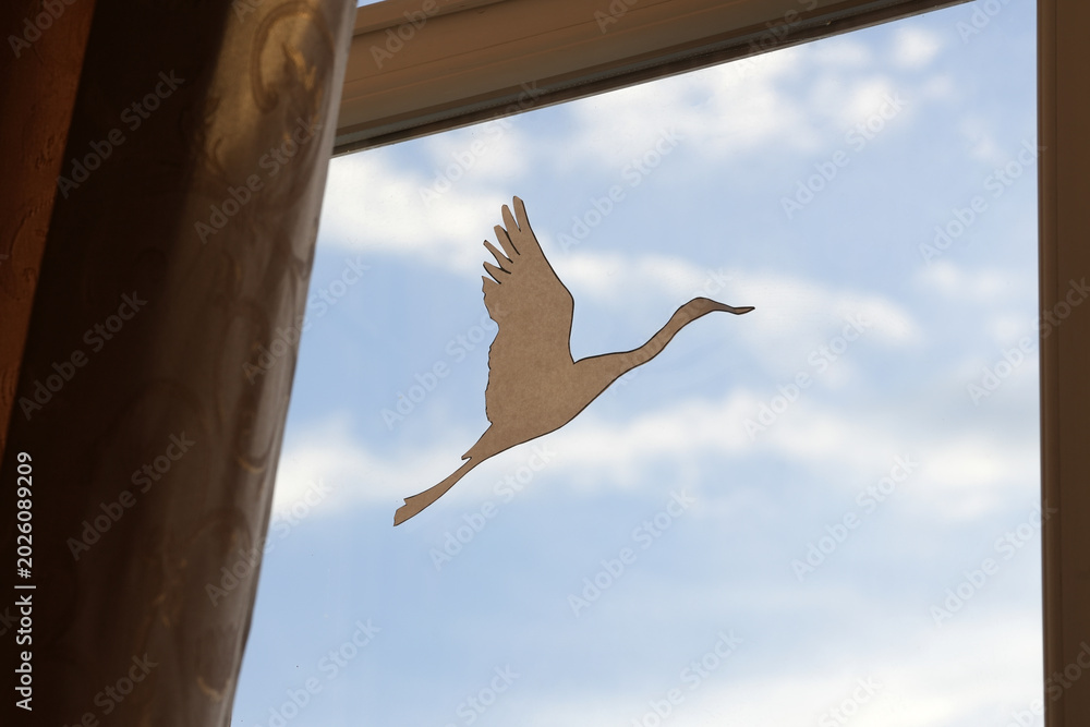 Naklejka premium Paper cutout of a flying crane positioned against a window with blue sky and clouds visible, creating a serene indoor atmosphere