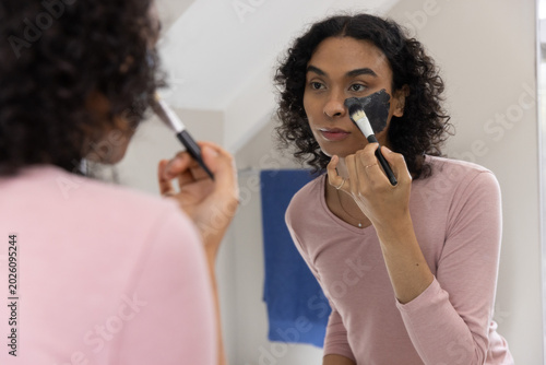 Non-binary adult applying dark face mask with flat brush at bathroom mirror wearing pink top