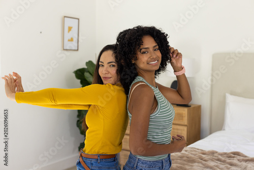 Diverse female friends wearing jeans standing back-to-back in bedroom, stretching near bed lamp