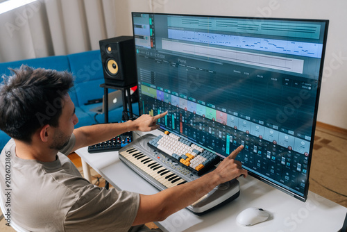 Tableau sur toile Rear view of sound engineer male creating new music in home studio, arranging tracks and mixing audio levels with midi keyboard and software on a large display, showcasing modern music production