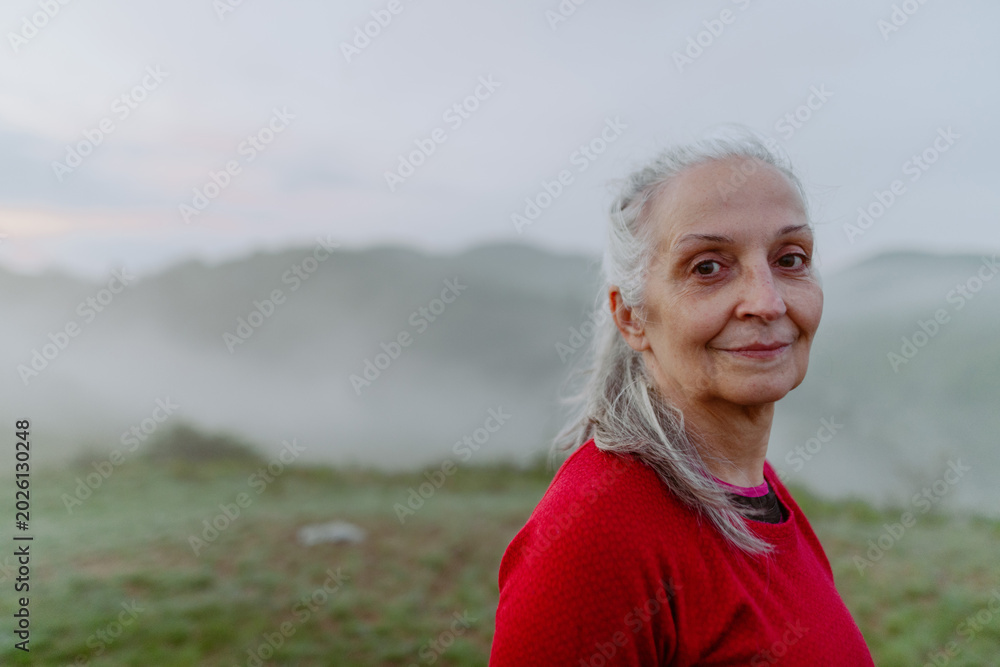 Fototapeta premium Senior woman hiking in nature on early morning with fog and mountains in background.