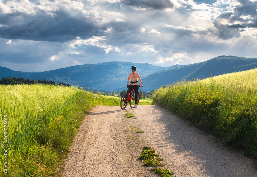 Obraz premium Female cyclist with mountain bike standing on dirt road in Slovenia countryside in summer. Woman looking at beautiful valley and mountains under dramatic stormy sky. Summer outdoor activity
