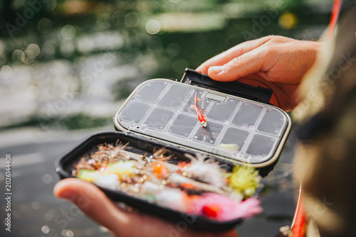 Young fisherman fishing on lake or river. Picture of plastic lures inside box for fishing time. Guy holding it in hands and show on camera. Stand in water. Daylight.